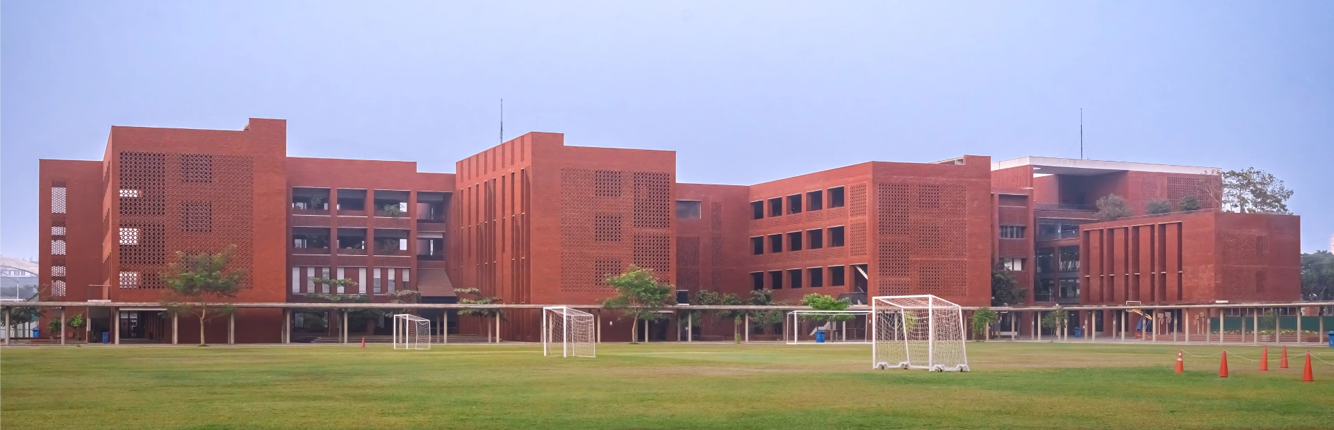 A wide view of the Aga Khan Academy Dhaka campus featuring its red-brick buildings, open green football fields, and clear blue sky.