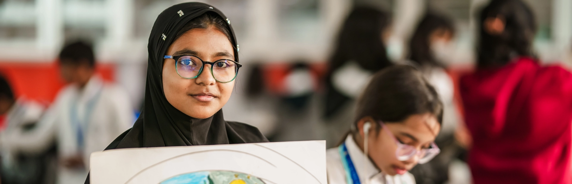 SlA student wearing glasses and a black headscarf holds up her colourful drawing featuring themes of nature, while another student works on art in the background.ider