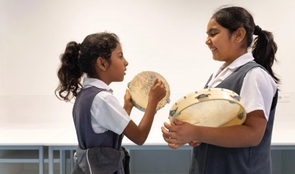 A teacher in a purple uniform holds up a colourful storybook while a group of young students sit at tables, listening and writing in the school library filled with books.