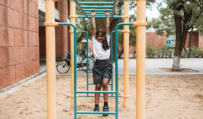 A teacher in a purple uniform holds up a colourful storybook while a group of young students sit at tables, listening and writing in the school library filled with books.