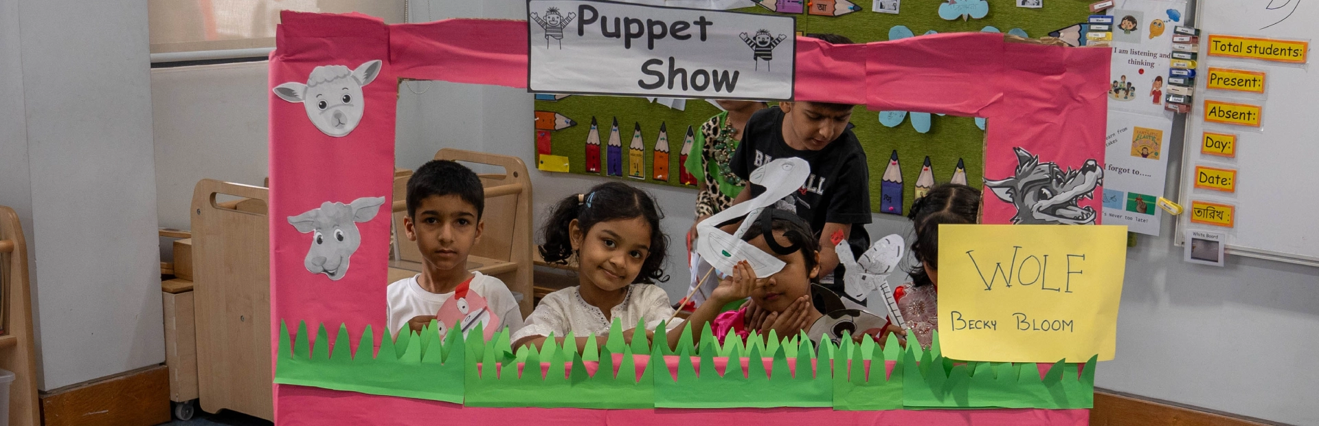 A teacher smiles as she assists two young students shaping clay letters during a classroom activity, with colourful learning materials and displays visible in the background