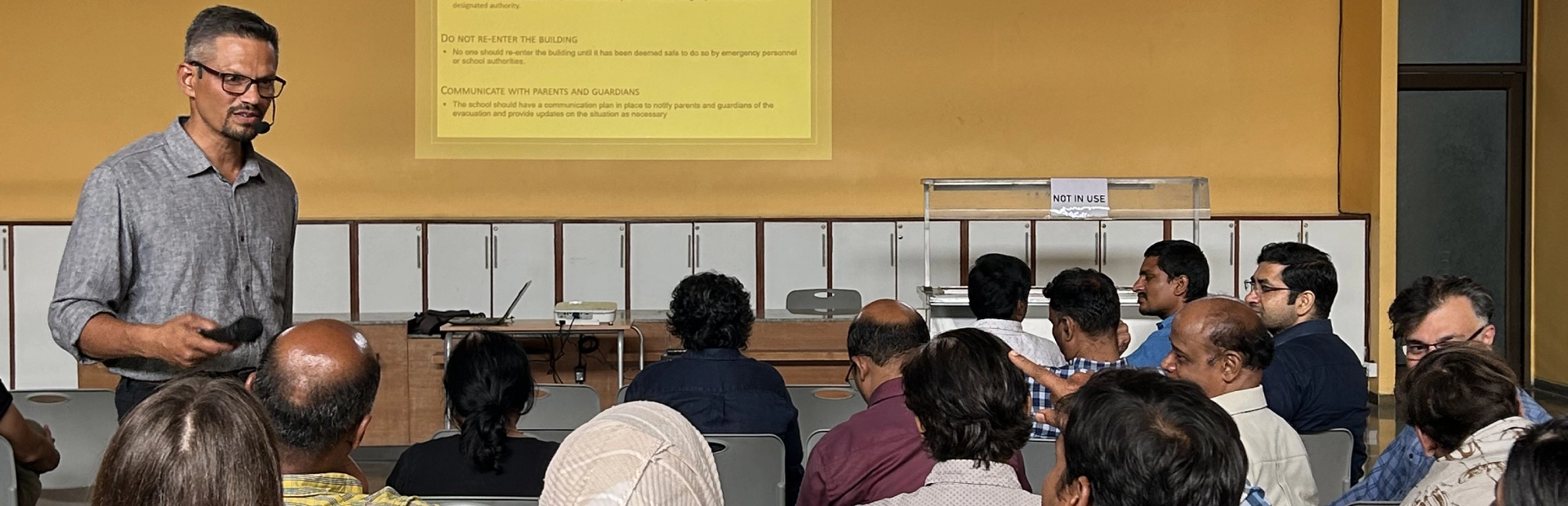 A diverse group of teachers sits closely together in a bright meeting room, listening attentively to a presentation. The participants appear engaged, reflecting a collaborative professional development atmosphere.