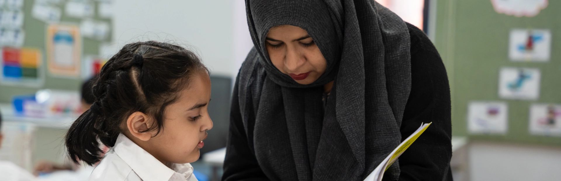 A teacher assists a young student in uniform inside a classroom at the Aga Khan Academy Dhaka.