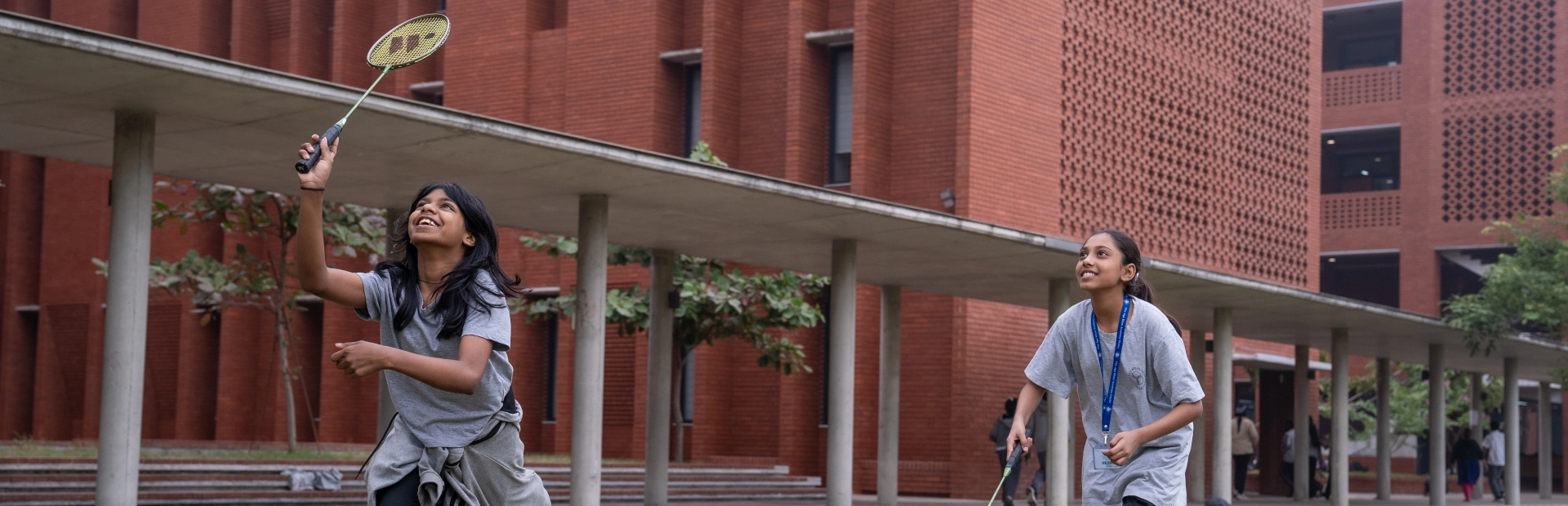 Two students play badminton on the school field, smiling as they enjoy the game against the backdrop of the red-brick campus.