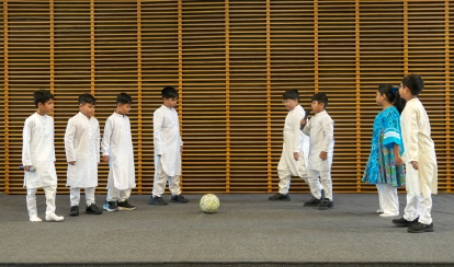 A group of young students dressed in traditional outfits stand on stage facing each other with a football between them, performing a drama in front of a wooden-panel backdrop.