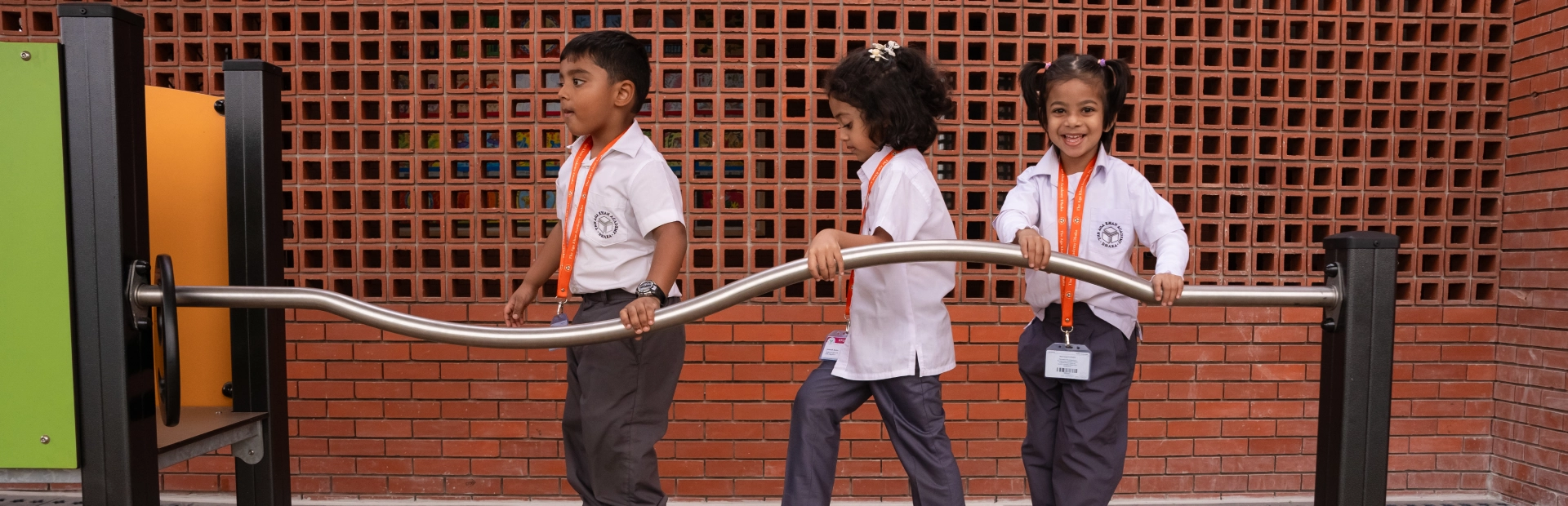 Three young students in school uniforms play on outdoor balancing equipment at the Aga Khan Academy Dhaka, smiling and enjoying their time together.