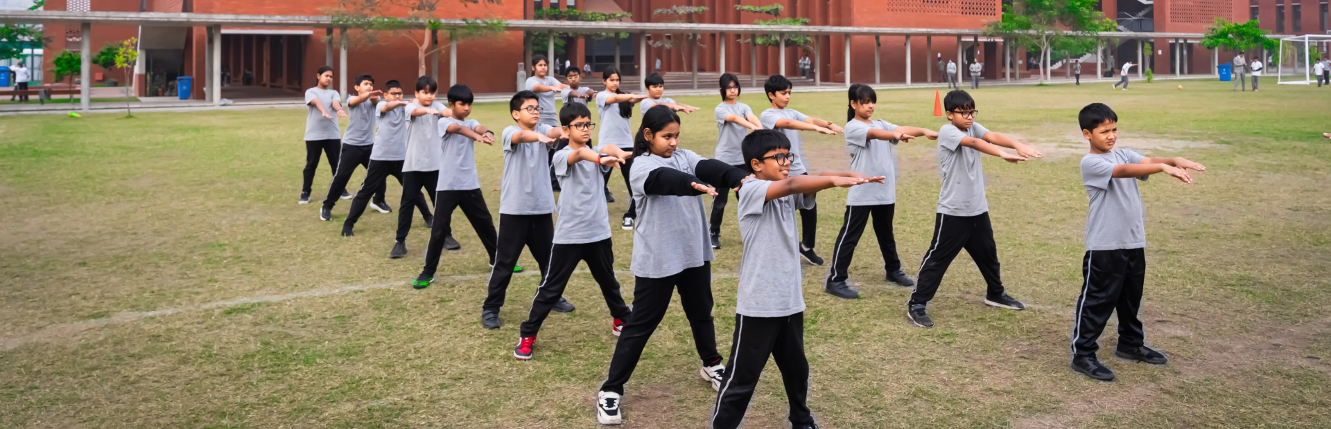 A group of students stand in formation on the school field performing morning exercises in front of the red-brick academic buildings under a clear sky.