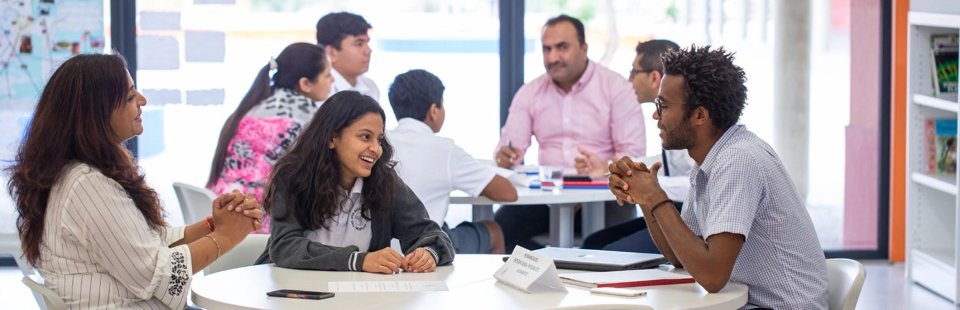 A teacher wearing a blue headscarf and red patterned outfit works with four Senior School students around a table, all engaged in discussion while looking at a laptop screen. The students, dressed in white uniforms with green lanyards, appear focused and collaborative in a modern classroom setting.