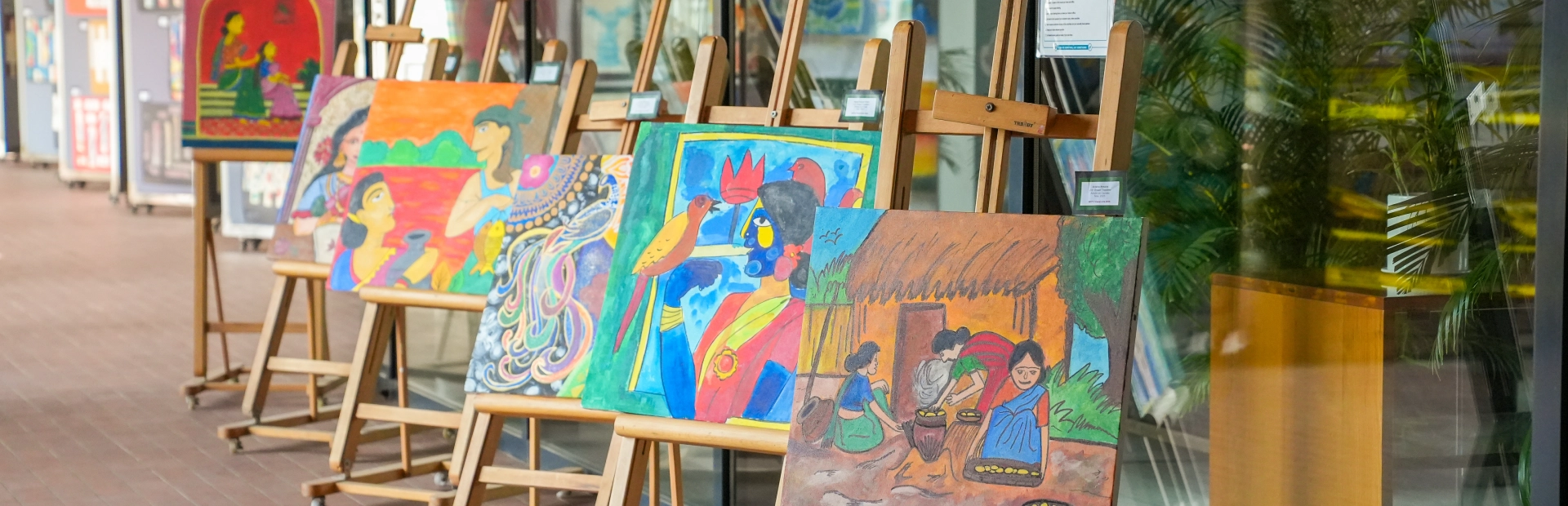 A row of colorful student paintings displayed on wooden easels along a corridor, featuring vibrant illustrations of people, culture and everyday life.