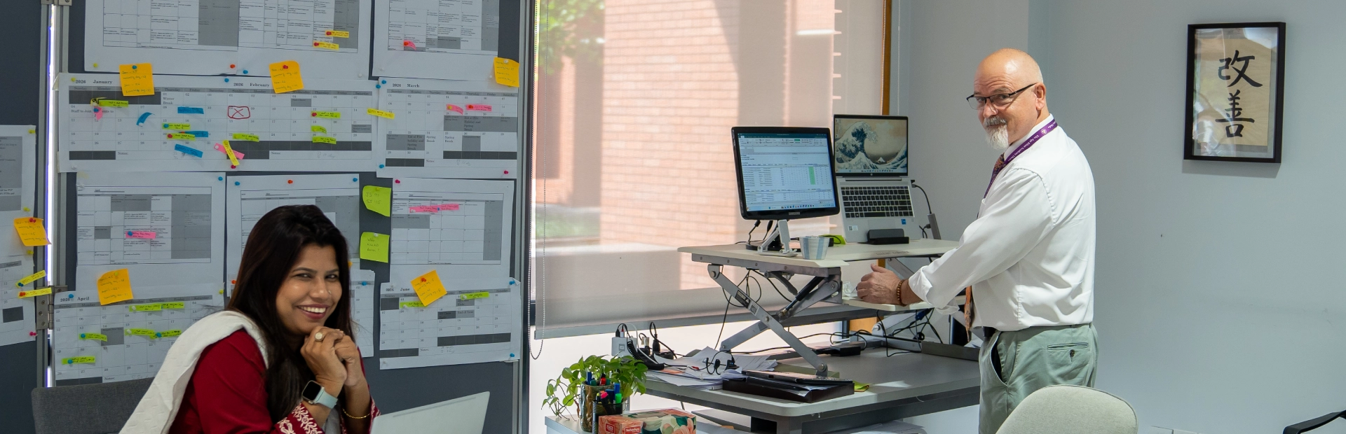 A woman sits at a desk smiling at the camera while a colleague works at a standing desk with multiple computer screens, in an office filled with colourful sticky notes and charts.